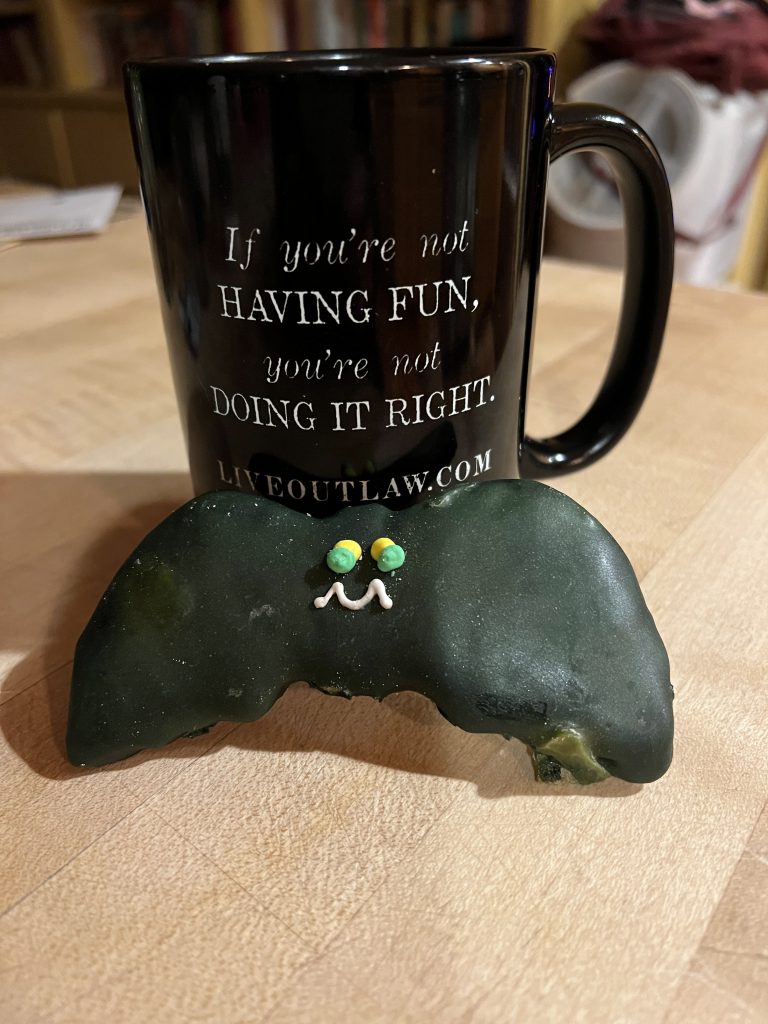 Tall, black mug with white lettering: If you're not having fun, you're not doing it right. Liveoutlaw.com. A black iced bat sugar cookie is propped against it on a wood kitchen counter.